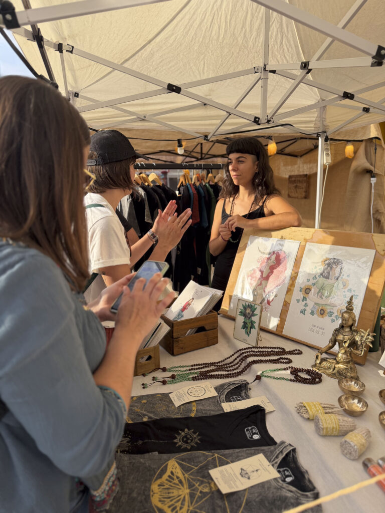 Visitants en una parada d'il·lustracions, maletes i joies artesanals al mercat de la Festa de la Malvaella 2026